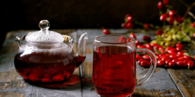Rose hip berries herbal tea in glass teapot Cup of rose hip berries herbal tea and glass teapot standing on old wooden plank table with wild autumn berries around. Winter hot beverage.